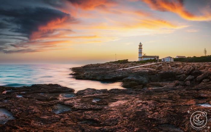 Photograph of Cap Salines Lighthouse in Mallorca at sunset, with warm sky and calm sea.