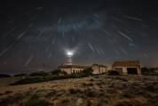 Cap de ses Salines lighthouse illuminated under a meteor shower during the Geminids, with radial trails in the night sky