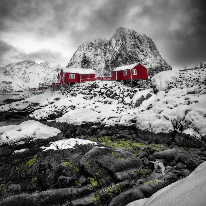 Winter landscape in Hamnøy, Norway, with rocks covered in green moss and red cabins contrasting with the snow and grey sky