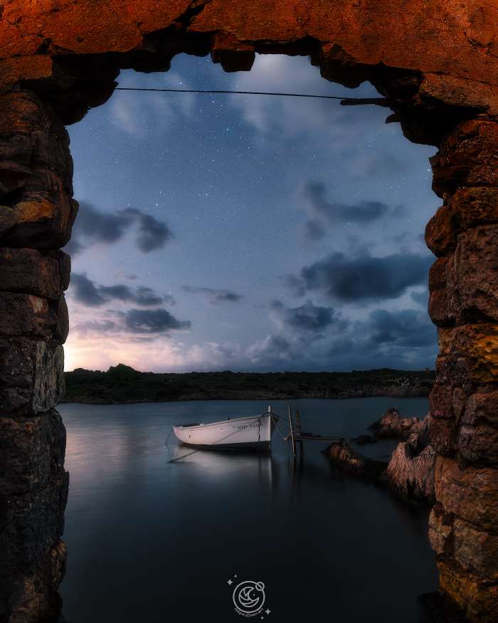 Llaüt amarrat al port de Sanitja, Menorca, vist des d’una caseta de pedra sota el cel estrellat